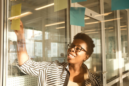 Black woman in glasses standing in modern office, looking at sticky notes stuck on glass wall and...
