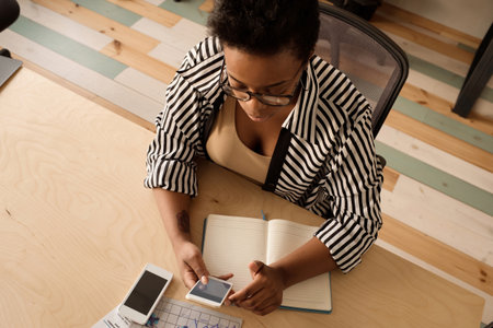 Stylish African American woman in striped blouse checking social media on smartphone while sitting at table in modern officeの写真素材