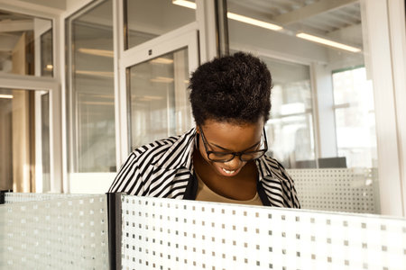 Black woman in striped blouse standing by partition wall in modern office, looking down and smiling