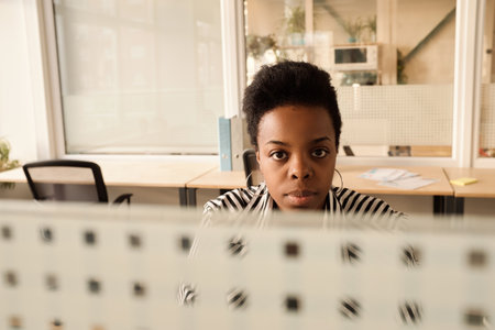 Portrait of African American woman sitting at table in modern office with divider walls and looking at cameraの写真素材