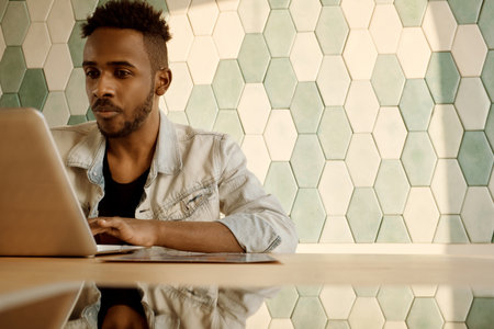 Young attractive black man in denim jacket sitting at table and working on laptop in room illuminated with bright sunlightの写真素材