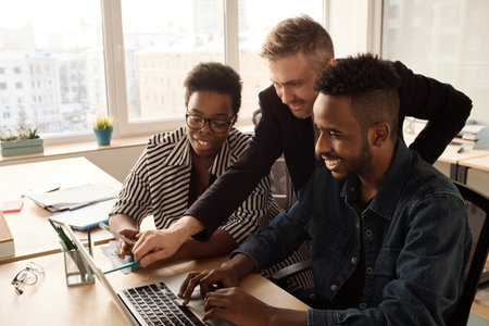 Multiethnic team of young managers looking at laptop, discussing new project and smiling in bright modern officeの写真素材