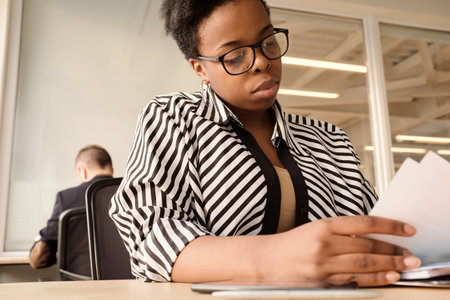 Low angle view of stylish black woman in striped blouse and glasses sorting documents while sitting at table in modern coworking spaceの写真素材