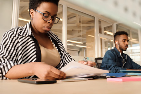 Low angle view of black woman working with papers in modern office, young man in earphones using computer besideの写真素材