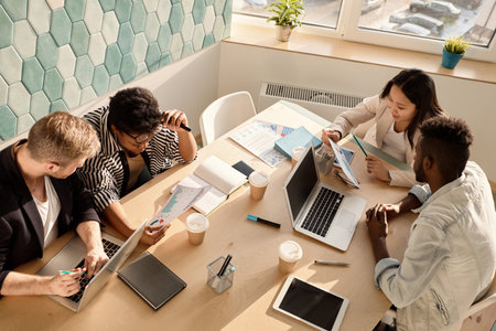 Multiethnic team of young businesspeople sitting at table in conference room and discussing new...
