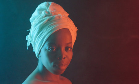 Headshot portrait of young attractive African woman in white turban with naked shoulders smiling at camera on dark background, among fogの写真素材