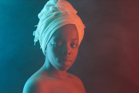 Close-up fashion portrait of young attractive Black woman in headwrap with naked shoulders looking away thoughtfully on dark backgroundの写真素材