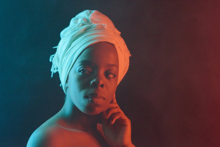 Waist-up portrait of young pretty African woman in white headwrap with bare shoulders looking aside thoughtfully on dark backgroundの写真素材