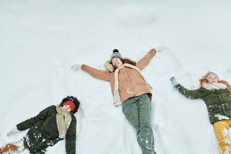 Caucasian woman lying in snow with two Caucasian children making snow angels, all smiling and looking up, winter clothing visible, outdoor winter activity, family bonding momentの写真素材