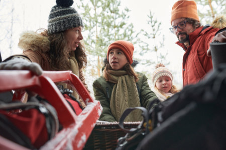 Caucasian family preparing sled outdoors in winter forest, all wearing warm clothing and hats, interacting togetherの写真素材