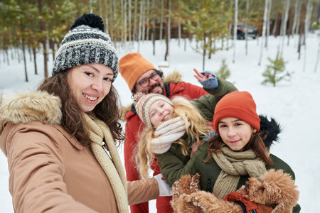 Caucasian woman taking selfie with Caucasian man and Caucasian siblings outdoors in snowy forest, all smiling and wearing winter hats, holding small dogの写真素材