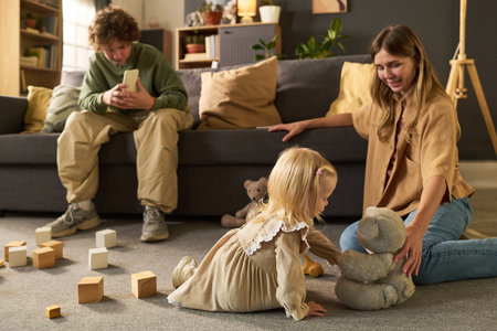 Caucasian young woman sitting on floor smiling at Caucasian child girl playing with teddy bear, while Caucasian boy sitting on sofa watching and holding smartphone in living roomの写真素材