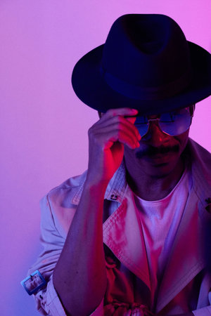 Portrait of African American man in trendy coat and sunglasses pulling down fedora hat over his face against purple backgroundの写真素材