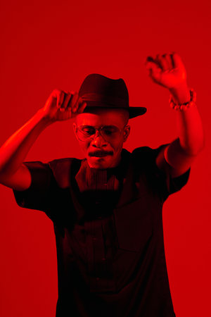 Portrait of stylish African American man with moustache in black clothes and fedora hat dancing in red neon lightingの写真素材