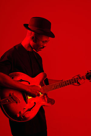 Portrait of stylish African American man in black clothes and fedora hat playing the guitar in red neon lightingの写真素材
