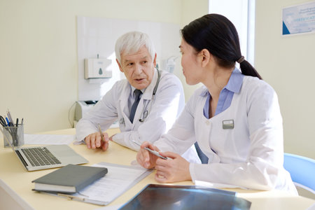 Professional mature physician and his young female assistant analyzing results of MRI scan in consulting room after examining radiographs of patientの写真素材