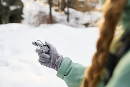 Caucasian young adult woman holding compass in gloved hand while standing outdoors in snowy landscape, long braided hair visible, navigating in winter environmentの写真素材