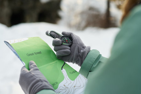 Caucasian young adult woman holding map and compass navigating snowy forest terrain, wearing gloves and outdoor jacket, focusing on route planning during winter adventureの写真素材