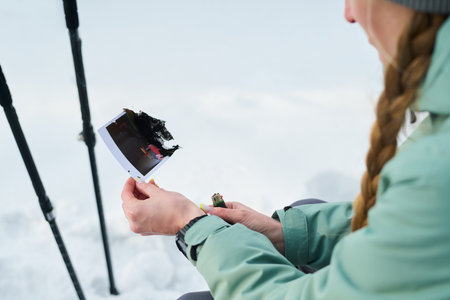 Young adult Caucasian woman holding partially burned instant photo outdoors, sitting in snow with trekking poles nearby, focusing on damaged photograph in handsの写真素材