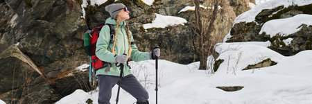Caucasian young adult woman hiking in snowy mountain landscape carrying backpack and trekking poles,...