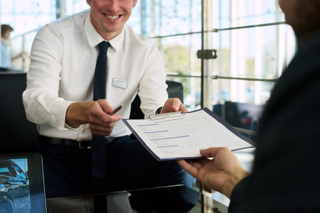 Caucasian young adult man smiling while handing contract and pen to middle aged man at car...