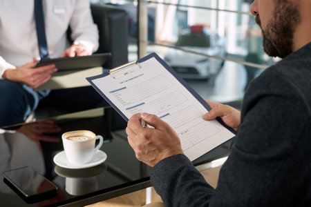 Middle aged Caucasian man holding pen and filling out car sale application form on clipboard while sitting at dealership tableの写真素材