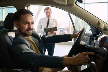Middle aged Caucasian man sitting in driver seat of car testing dashboard controls while young Caucasian man standing outsideの写真素材