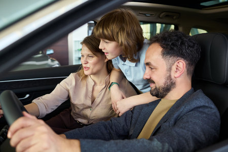 Middle aged woman sitting in driver seat showing car dashboard to middle aged man and child leaning forward inside car dealership showroomの写真素材