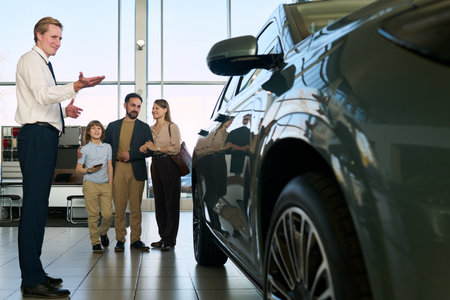 Caucasian male car salesman presenting vehicle to Middle aged Caucasian man, Middle aged Caucasian woman, and child boy standing together inside modern car dealership showroomの写真素材