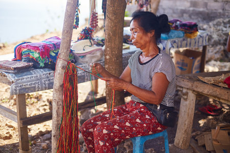 Bali, Indonesia, November 18, 2015. Middle aged Asian woman sitting outdoors weaving colorful threads by hand, surrounded by handmade crafts and textiles displayed on wooden tablesのeditorial素材