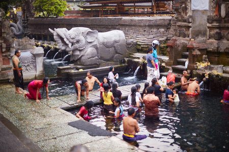 Bali, Indonesia, November 18, 2015. Group of men, women, and children participating in traditional purification ritual in temple pool, standing and kneeling in water near stone elephant fountainのeditorial素材