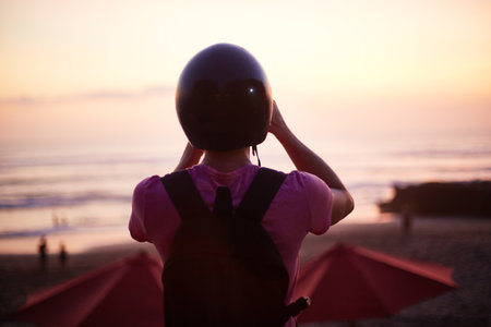 Woman wearing black helmet standing on beach at sunset, holding helmet with both hands, backpack...