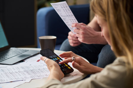 Caucasian young woman using smartphone calculator while reviewing tax documents with Caucasian man,...
