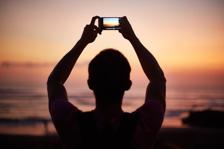 Man holding smartphone above head capturing sunset over ocean, standing outdoors with arms raised, silhouetted against colorful sky during eveningの写真素材