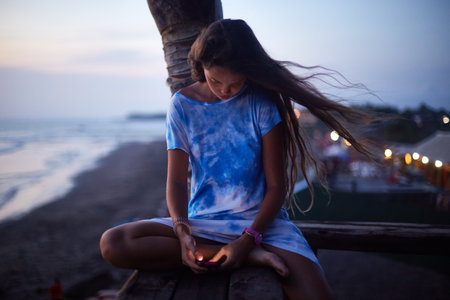 Young woman sitting cross legged on wooden railing near beach at sunset, looking down at smartphone in hands, long hair blowing in wind, relaxed outdoor settingの写真素材
