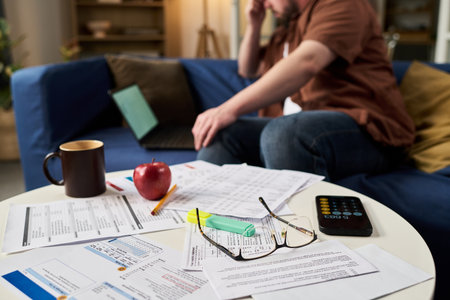 Caucasian young adult man sitting on sofa using laptop and talking on phone while reviewing tax...