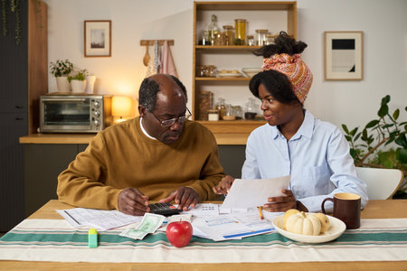 Middle aged Black man using calculator and reviewing financial documents with young Black woman sitting at table, both focusing on paying taxes, paperwork and money visible on surfaceの写真素材