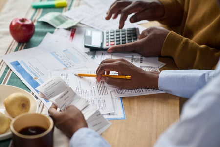 Closeup of Black man and Black woman calculating taxes together at table, reviewing receipts and financial documents with calculator, hands visible holding pencil and paper, coffee and fruit nearbyの写真素材