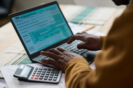 Middle aged Black man using laptop and calculator while filling out tax forms at desk, hands typing on keyboard with tax documents and financial paperwork spread out on tableの写真素材