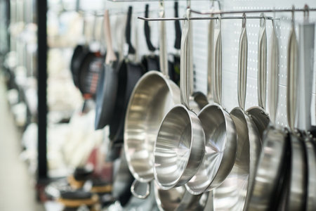 Stainless steel frying pans and saucepans hanging on hooks in kitchenware store, cookware displayed in organized row with blurred background showing additional utensils and equipmentの写真素材