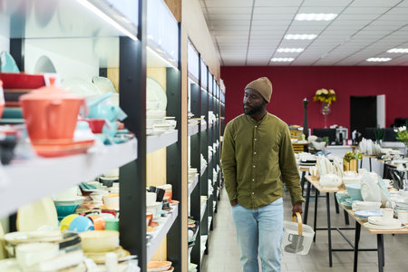 Black young adult man walking through store aisle holding shopping basket, browsing shelves filled with various kitchenware and tableware, looking at displayed productsの写真素材