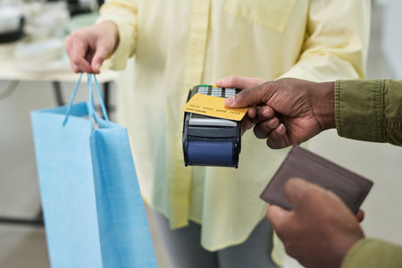 Caucasian woman handing shopping bag while Black man making payment with credit card using point of sale terminal, both adults engaged in retail transaction, close up on handsの写真素材