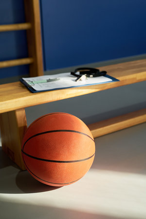Basketball resting on gym floor near wooden bench with clipboard and whistle placed on top, suggesting preparation for sports training or coaching session in athletic facilityの写真素材