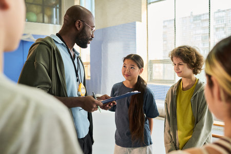 Black man coach holding clipboard talking to diverse group of teenagers including Asian girl and...