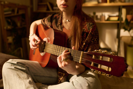 Young adult Caucasian woman sitting cross legged playing acoustic guitar in cozy indoor setting, focusing on hand positioning and finger placement on fretboardの写真素材