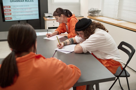 Three young adult female inmates sitting at table writing on worksheets during prison education class, focused on completing assignmentsの写真素材