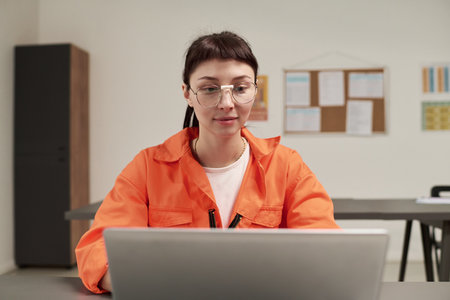 Caucasian young adult woman wearing glasses sitting at desk using laptop computer in classroom setting participating in prison education program with focus on learningの写真素材