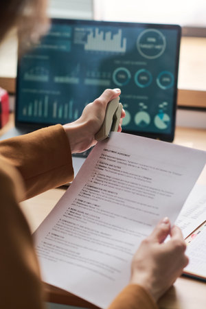 Young adult woman holding employment agreement document while stapling papers at desk, computer monitor with business analytics and charts visible in backgroundの写真素材