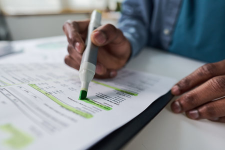 Black man highlighting financial data on printed document with green marker, focusing on analyzing numbers and making notes, closeup of hands working at desk in office settingの写真素材