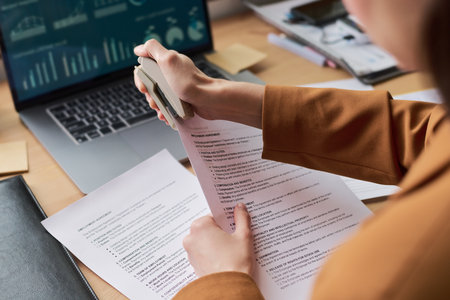 Caucasian young adult woman stapling contract documents at desk, holding papers in hands, working in office with laptop displaying financial charts, performing business taskの写真素材